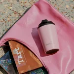 A pink reusable cup with a black lid rests on a pink bag, beside decorative items on a textured surface.