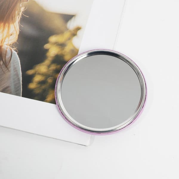 A circular tinplate pocket mirror with a silver reflective surface and a pink border, placed next to a photograph. A circular tinplate pocket mirror with a silver reflective surface and a pink border, placed next to a photograph.