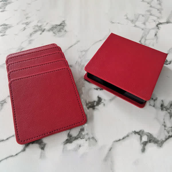 Four red PU leather coasters stacked beside a matching storage case on a marble surface. Four red PU leather coasters stacked beside a matching storage case on a marble surface.