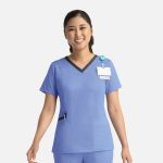 A nurse in a blue scrub top with a contrast V-neck and pocket details, wearing a badge and a smile.