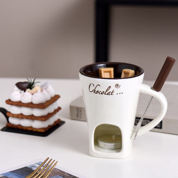 A chocolate fondue cup in cream and brown, featuring a tealight holder and a fondue fork. Includes a dessert in the background. A chocolate fondue cup in cream and brown, featuring a tealight holder and a fondue fork. Includes a dessert in the background.