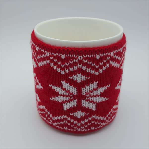 A knitted cup sleeve in red and white with a snowflake design wraps around a plain white mug. A knitted cup sleeve in red and white with a snowflake design wraps around a plain white mug.