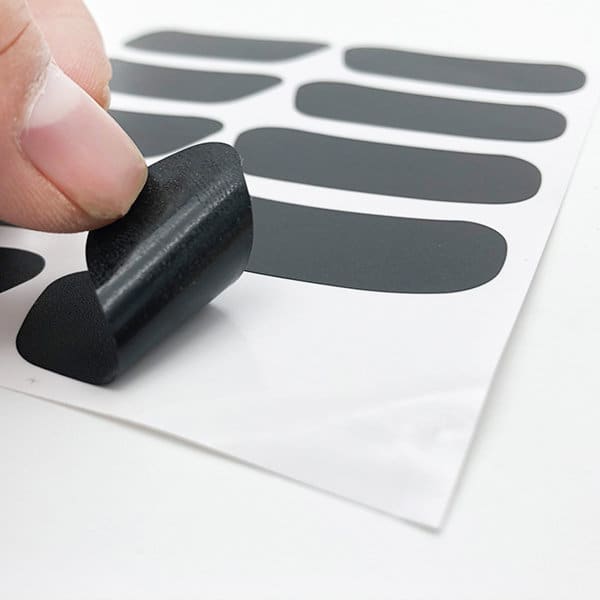 A hand lifts a black custom eye black sticker off a sheet, featuring multiple stickers in various sizes. A hand lifts a black custom eye black sticker off a sheet, featuring multiple stickers in various sizes.