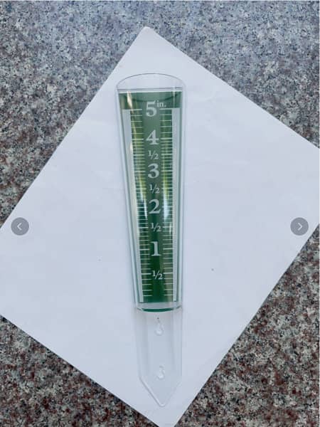 A clear, plastic magnifying rain gauge in green, displaying measurements from 0 to 5 inches on a flat surface. A clear, plastic magnifying rain gauge in green, displaying measurements from 0 to 5 inches on a flat surface.