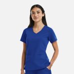 A woman stands wearing a blue curved V-neck top, featuring short sleeves and two front pockets. It has a logo on the sleeve.