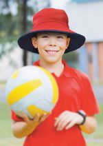 A red and navy reversible bucket hat is worn by a smiling child holding a yellow and white volleyball.