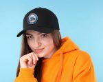 A woman wearing a black cotton twill cap with a buckle, paired with an orange hoodie. The cap features a logo.