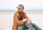 A man wearing an olive green cap sits on the beach, dressed in a brown jacket and grey jeans, with the ocean in the background.