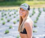 A woman with long blonde hair wears a grey and black trucker cap, smiling outdoors in a field of plants.