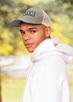 A man in a grey cap and white hoodie stands outdoors, showcasing a stylish look against a blurred natural background.