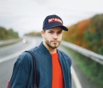 A man wearing a navy cap with a logo, a blue bomber jacket, and a red shirt, standing near a road with autumn trees.