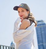 A woman wearing a black cap and white shirt poses outdoors, with a city skyline in the background.