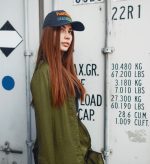 A woman with long brown hair wears a blue cap and a green jacket, standing against a shipping container background.