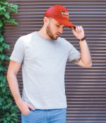 A man wearing a grey t-shirt and a red cap stands against a textured background with greenery. The cap features a logo.