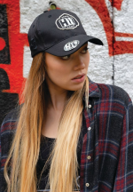 A black twill cap with a logo, worn by a model with long hair, standing in front of a graffiti background.
