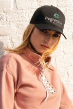 A black cotton cap featuring a logo, worn by a model in a pink top against a light brick background.