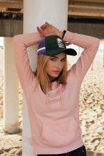A woman in a pink hoodie poses under a structure at the beach, wearing a green and purple trucker cap.