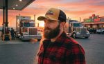 A man with a beard wears a tan and black trucker cap, standing beside trucks at a gas station during sunset.