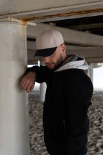 A man leans against a concrete pillar wearing a black jacket and a beige and black cap.