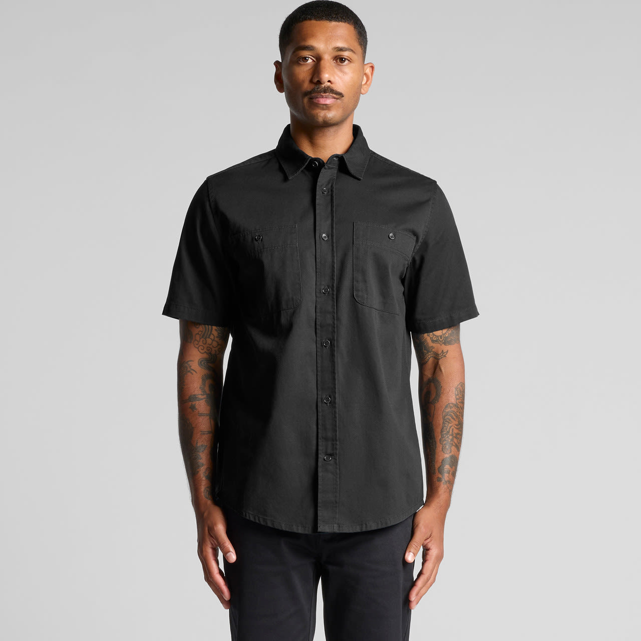 A short-sleeved black work shirt displayed on a model with tattoos, featuring two front pockets and a logo. A short-sleeved black work shirt displayed on a model with tattoos, featuring two front pockets and a logo.