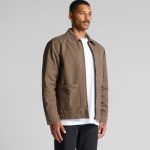 A brown canvas heavy jacket featuring two front pockets and a logo, worn by a model against a neutral backdrop.