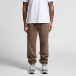 Brown track pants with a relaxed fit and an elastic waistband, paired with grey sneakers. The model wears a plain white t-shirt.