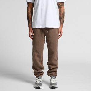 Brown track pants with a relaxed fit and an elastic waistband, paired with grey sneakers. The model wears a plain white t-shirt.
