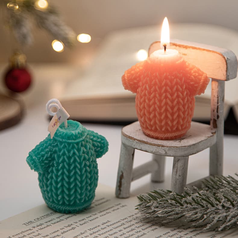 Two sweater-shaped candles in teal and orange, sitting on a small chair with soft lighting and a blurred background. Sweater Shape Candles - Image 1