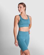 A woman poses in a teal sports cropped tank top and matching high-waisted shorts, featuring a logo.