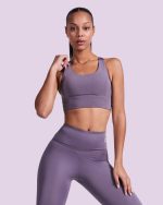 A woman poses in a purple cropped tank top and matching leggings, with a simple background. The top has a logo.