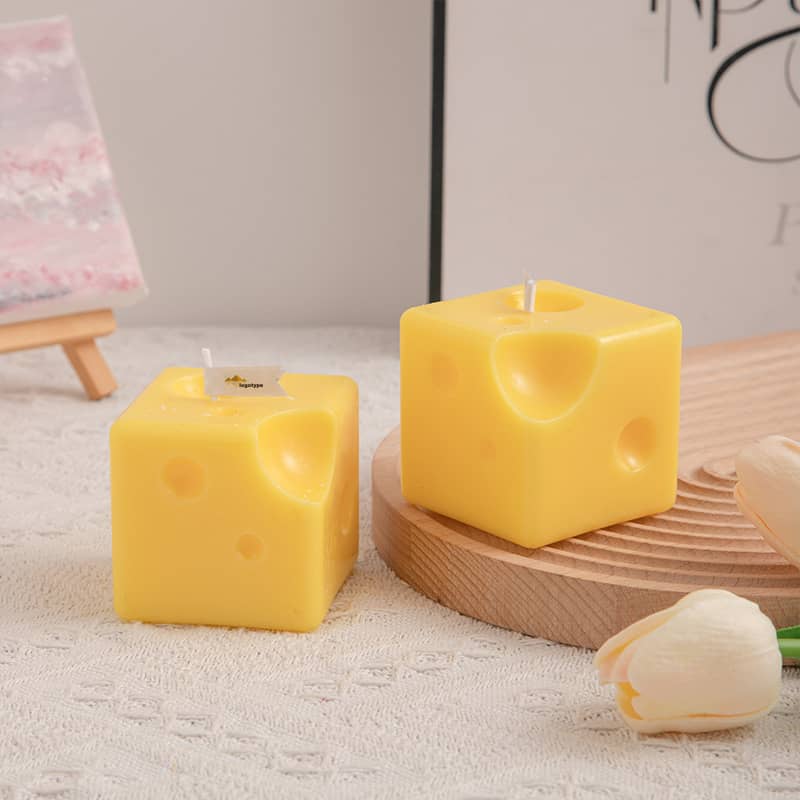 Square candles shaped like cheese, featuring a yellow colour with holes, displayed on a wooden plate with flowers nearby. Square Cheese Shape Candles - Image 1