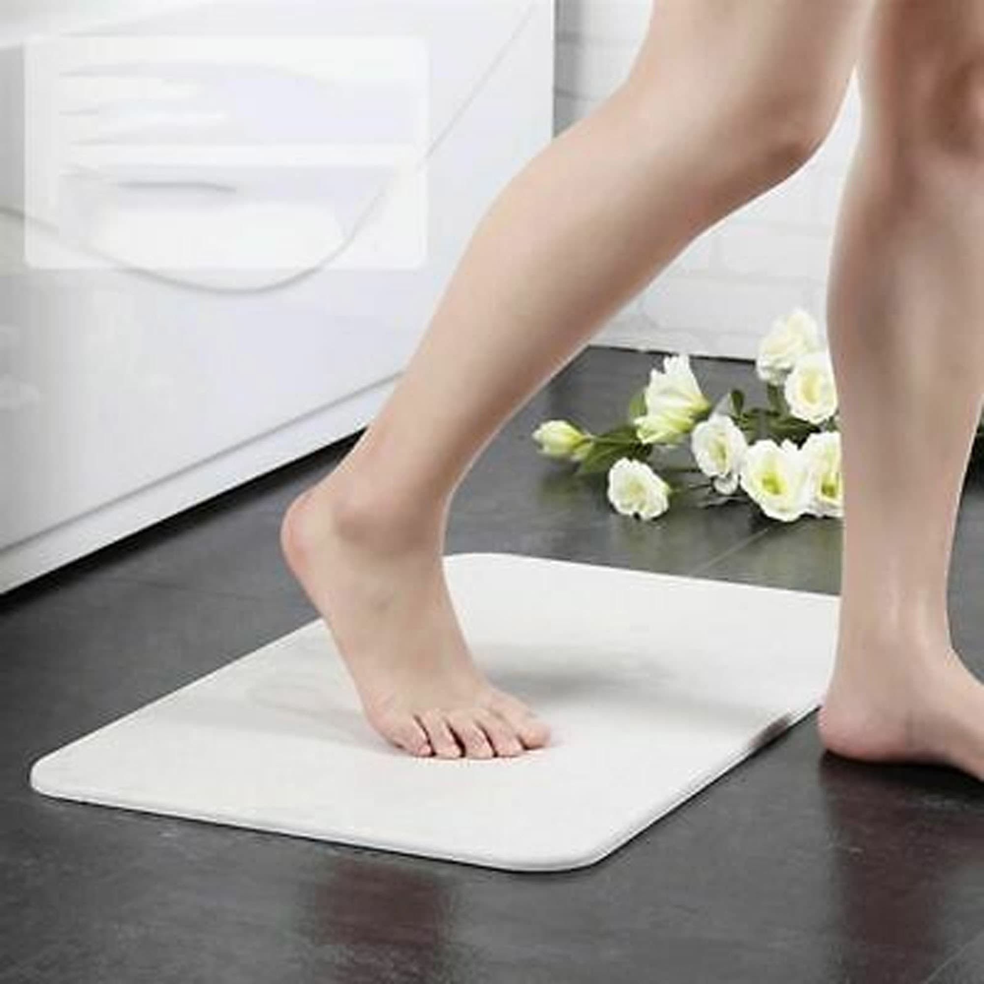 A soft white bath mat with a foot stepping onto it, placed on a tiled floor near flowers. It has a logo. Toyo I-Dried Bath Mat - Image 1