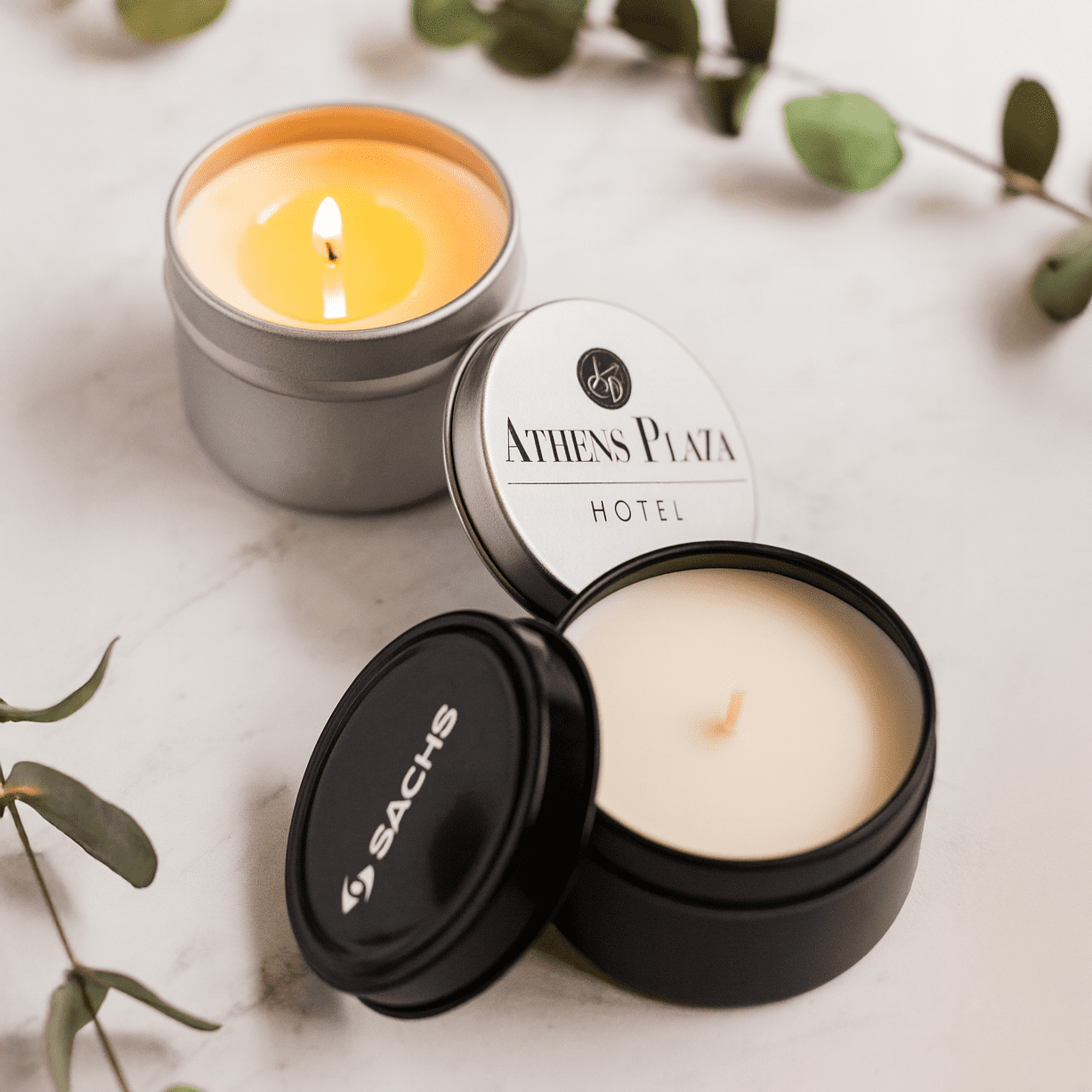 Three scented candles in silver and black containers, one with a lit wick, accompanied by green leaves. Vanilla Scented Candle - Image 1