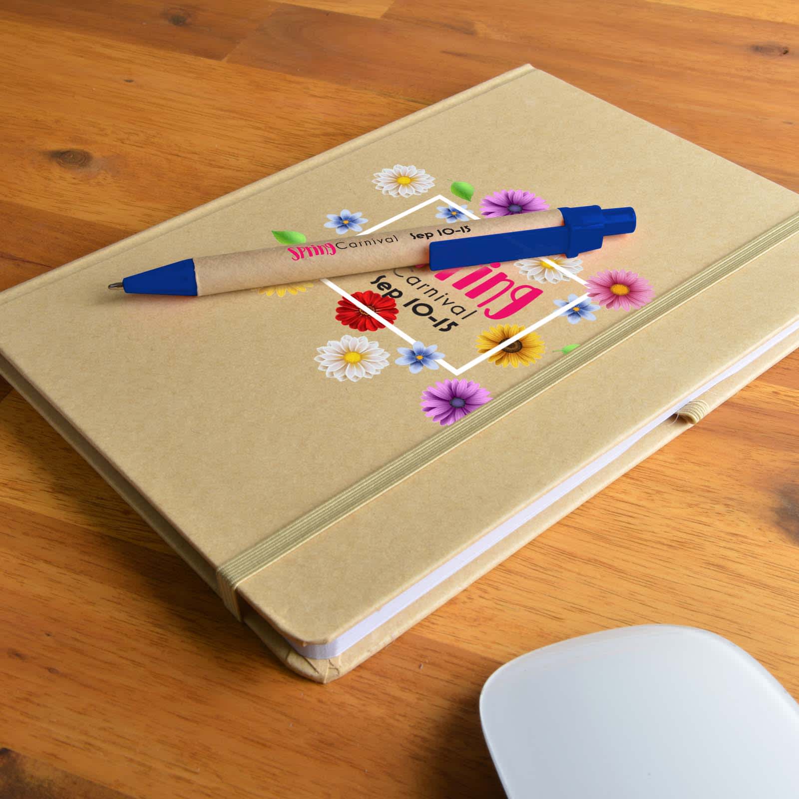A tan A5 Natura notebook with a floral design and a blue Matador pen on a wooden surface. Venture A5 Natura Notebook / Matador Pen - Image 1