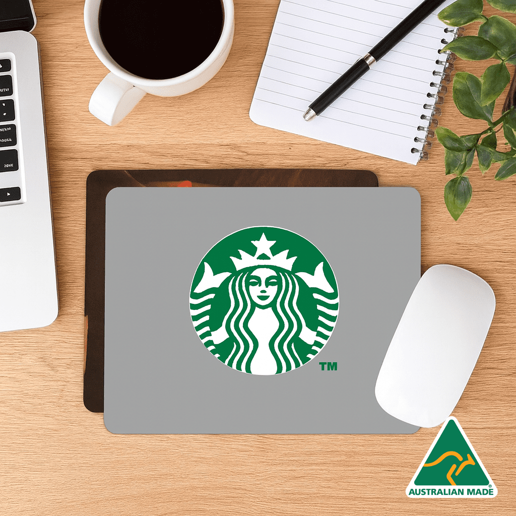 A grey mouse mat featuring a logo, placed on a wooden desk with a coffee cup, notebook, and plant. Deluxe Mouse Mat (230mm x 190mm) - Image 1