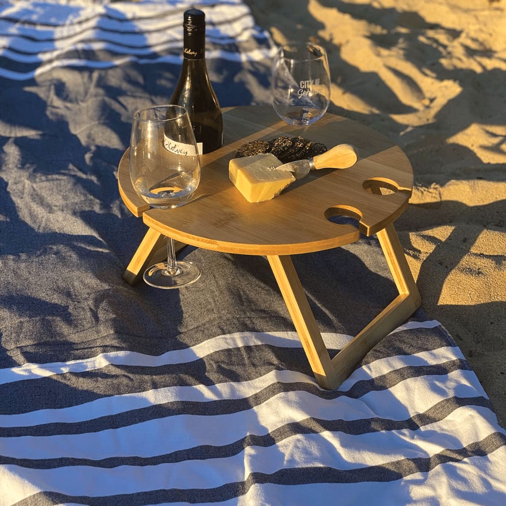 A round bamboo picnic table with drink holders, set on a striped blanket, accompanied by wine glasses and a cheese board. Trekk Picnic Table - Image 1