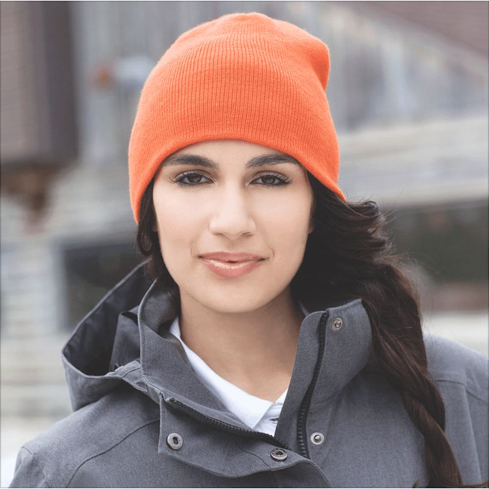 An orange knit beanie worn by a person with long dark hair, paired with a grey jacket. The beanie has a logo. Level Knit Beanie - Unisex - Image 1