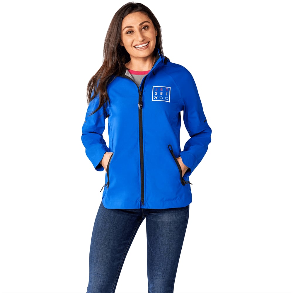 A woman wearing a blue softshell jacket with a logo, featuring a zip front and side pockets. Oracle Softshell Jacket - Womens - Image 1