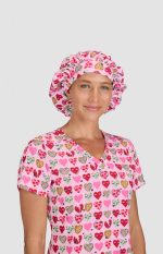 A pink bouffant cap with a heart print, worn by a model in a matching scrubs top with a heart design.