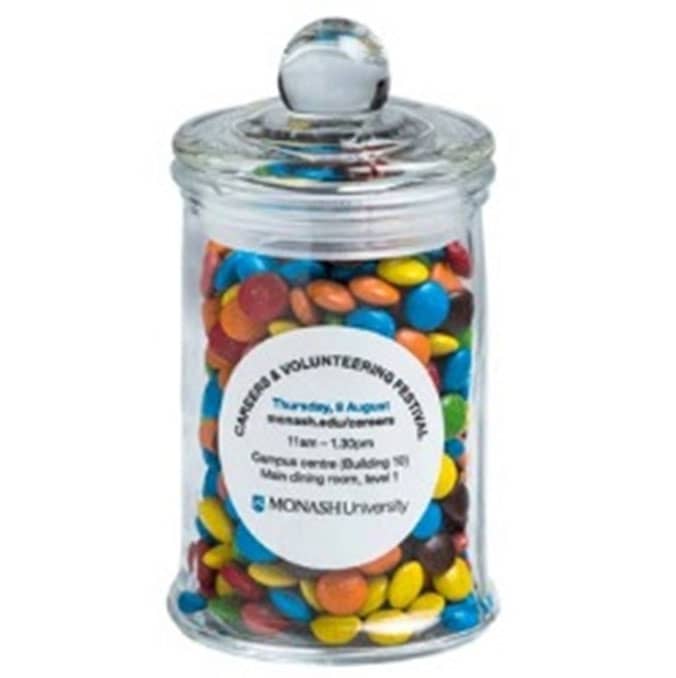 A small glass apothecary jar filled with colourful chocolate beans, featuring a round lid and a logo. Small Apothecary Jar with Mini M&Ms 115g - Image 1