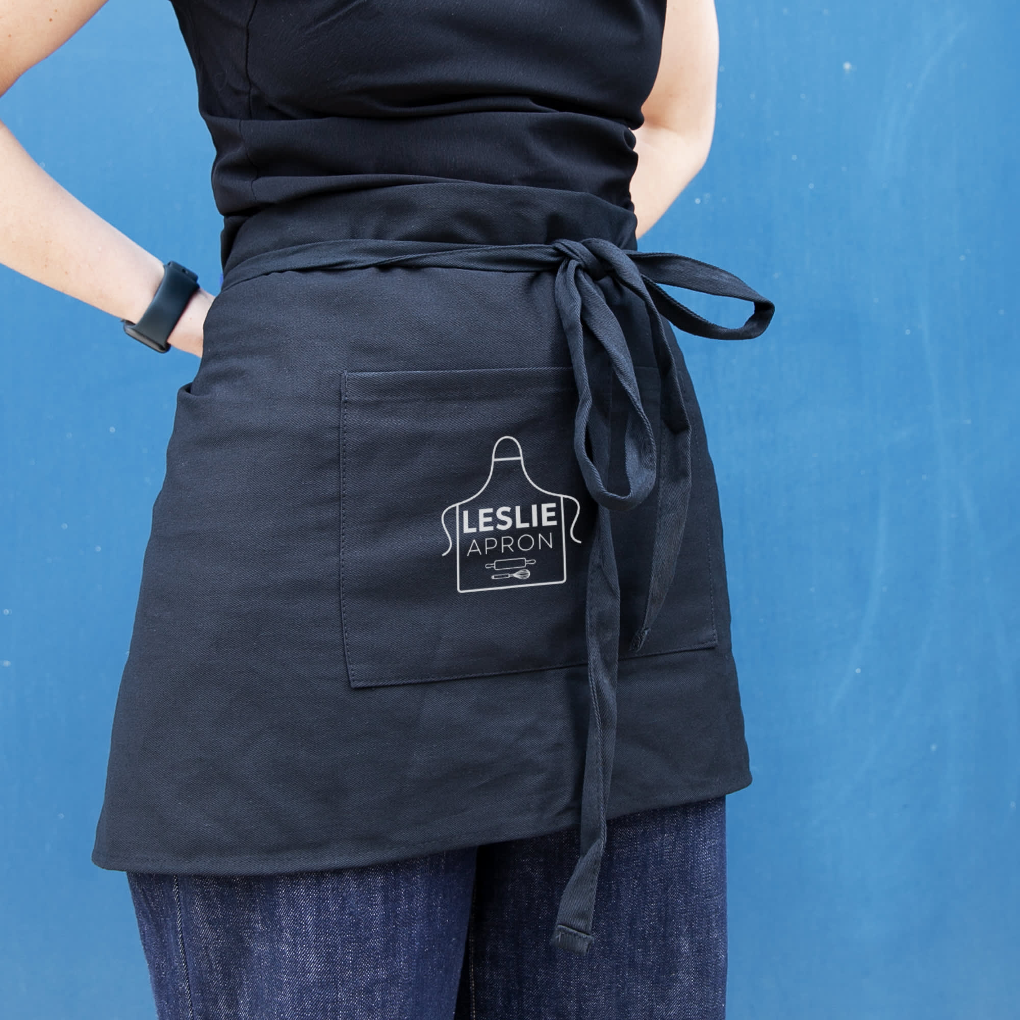 A black waist apron with a front pocket and a tied waist, held by a person against a blue background. Leslie Waist Apron - Image 1