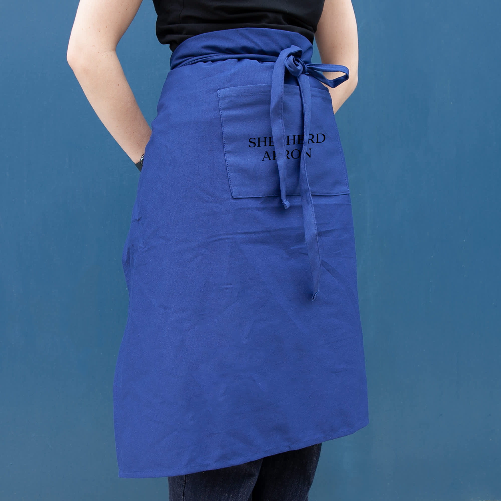 A blue long waist apron with a front pocket and tied at the waist, displayed against a blue background. Shepherd Long Waist Apron - Image 1
