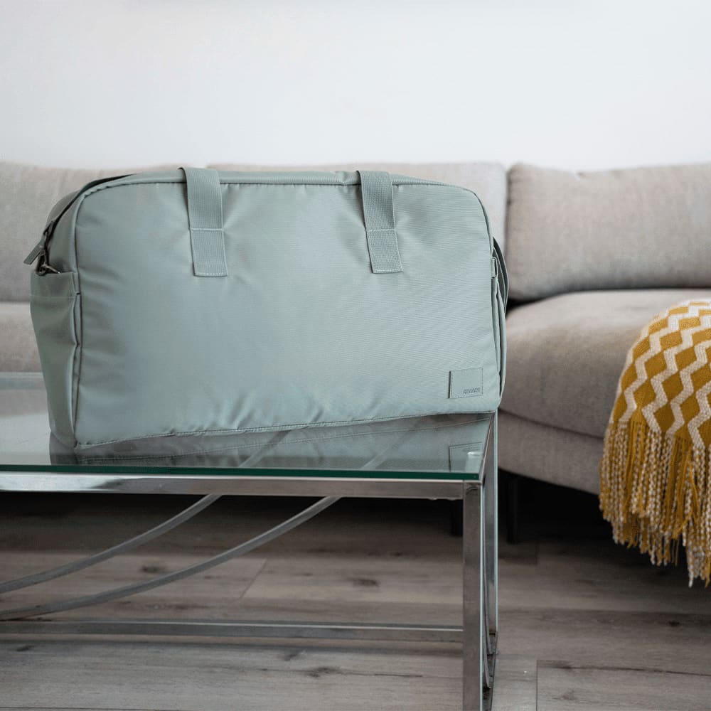 A green duffle bag sits on a glass table, with a sofa and blanket in the background. The bag has handles and a logo. American Tourister Urban Groove Duffel - Image 1