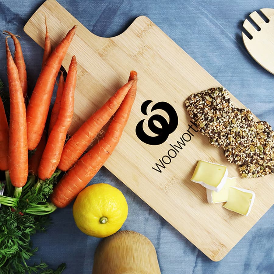 A bamboo chopping board featuring a logo, with fresh carrots, a lemon, and slices of cheese on display. Ozi Bamboo Paddle Board - Image 1