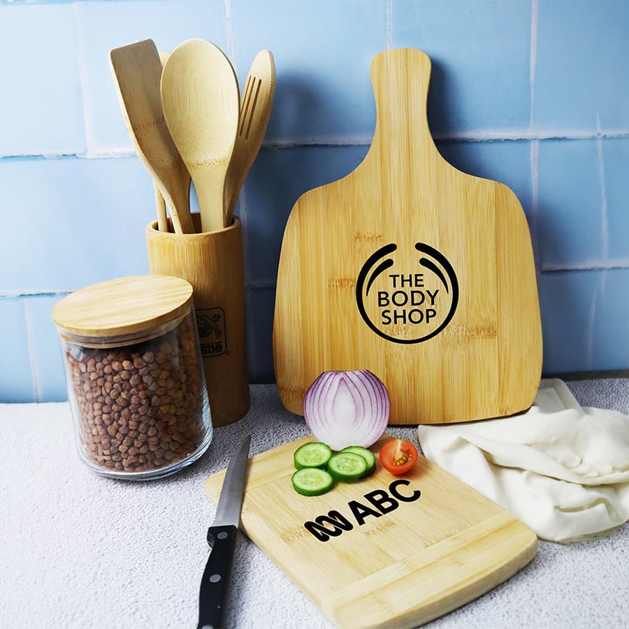A bamboo cutting board in natural colour, alongside fresh vegetables, utensils, and a jar of ingredients. Features a logo. Tiga Bamboo Cutting Board - Image 1