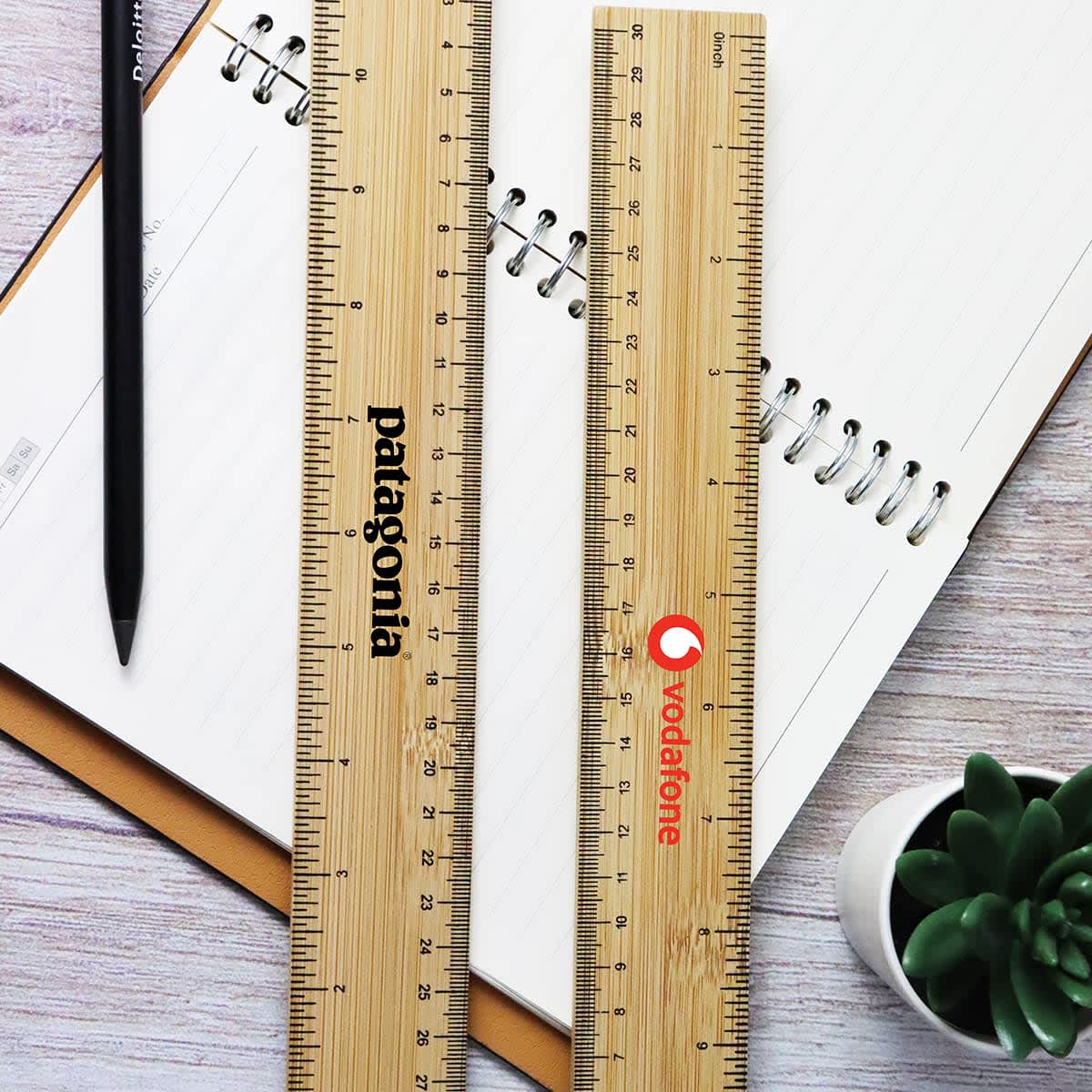 Two bamboo rulers, one with a logo, placed on a notebook with a pen and a small plant nearby. Apex Bamboo Ruler - Image 1