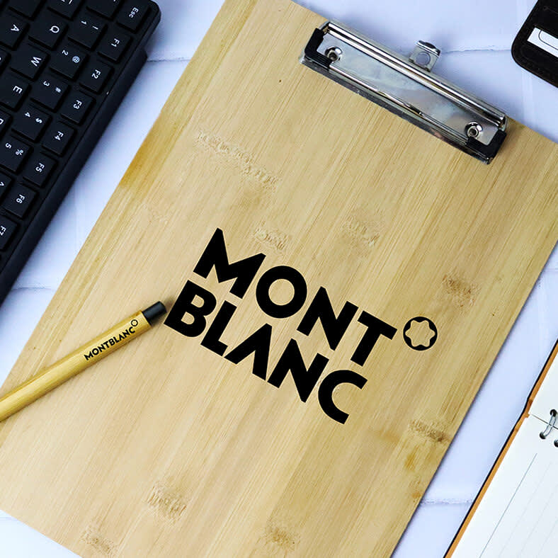 A bamboo clipboard with a dark logo, placed on a surface with a keyboard, pen, and notebook. Ecowriter Bamboo Clipboard - Image 1