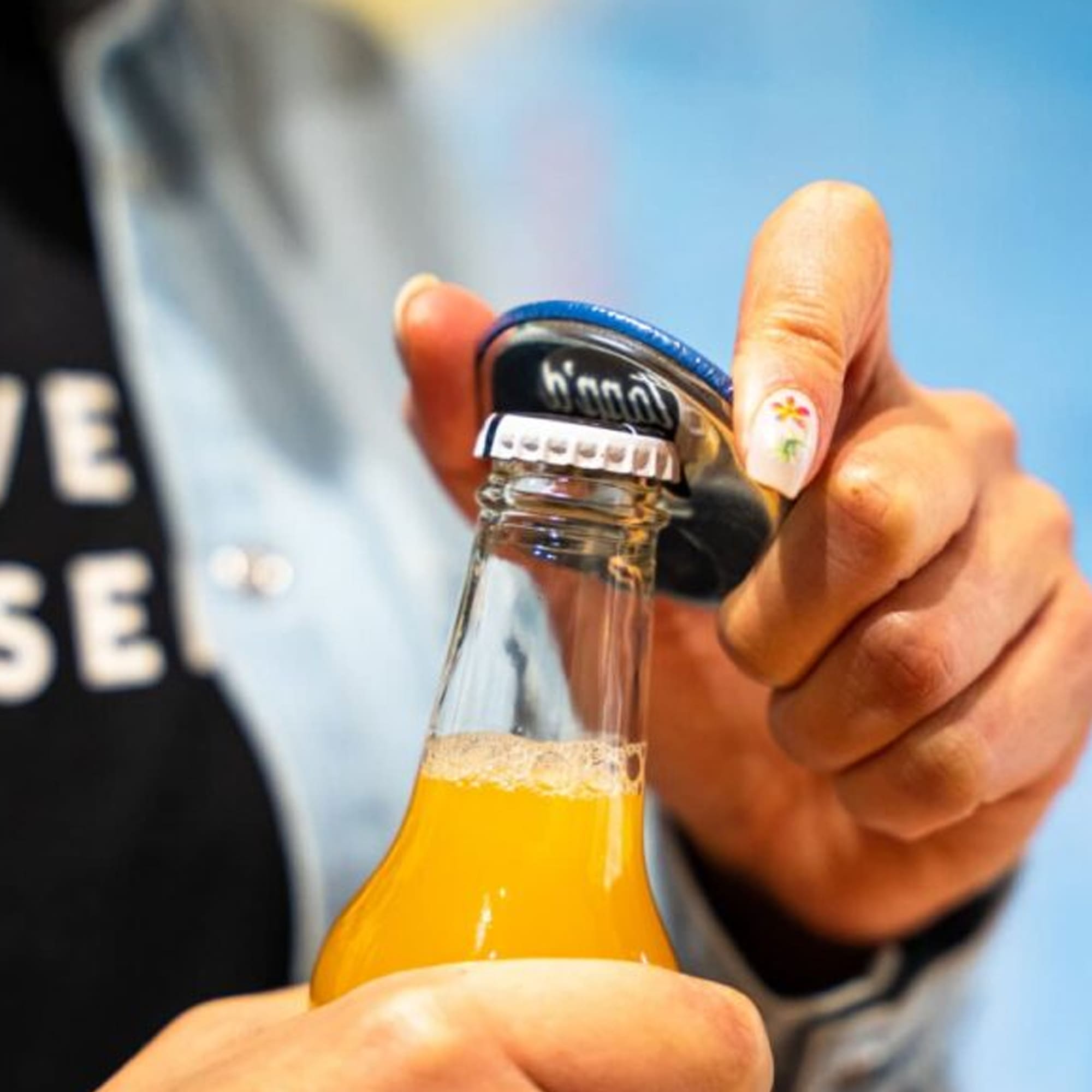 A drink bottle opener with a blue handle is being used to cap a glass bottle of yellow beverage. Button Badge Fridge Magnet Bottle Opener - Image 1