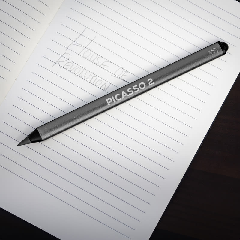 A grey stylus pen resting on an open notebook with a lined page. The pen features a logo and product name. Picasso 2 Eco Stylus Pen - Image 1