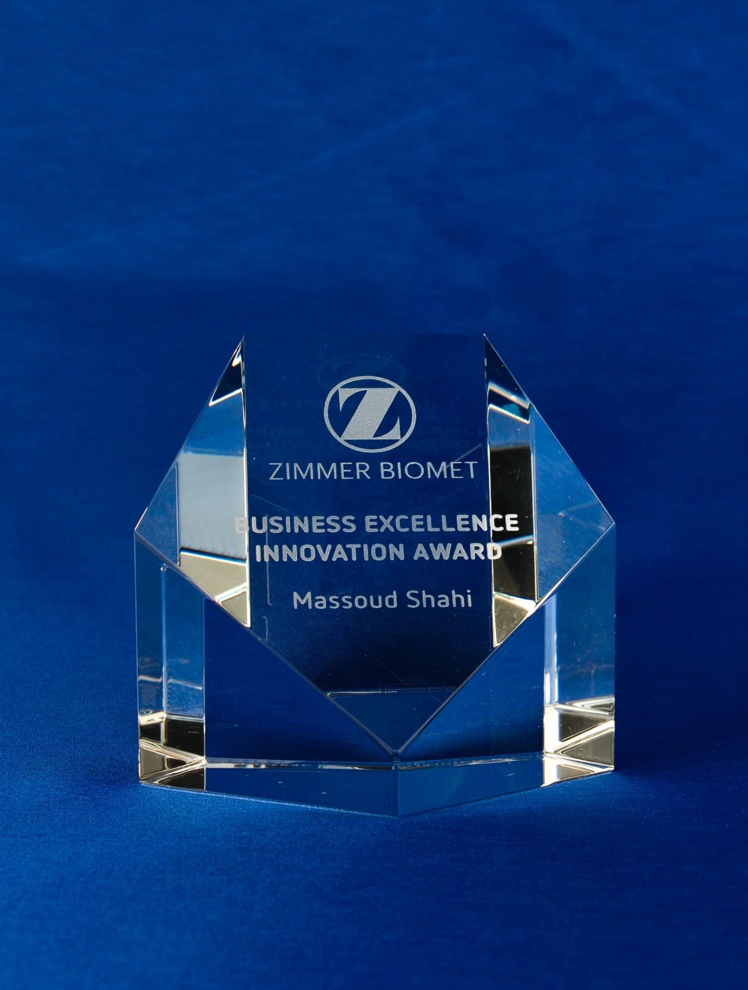 A clear crystal pentagon paperweight with an engraved award, displayed on a blue background. It has a logo and text. Sliced Crystal Pentagon Paperweight + Gift Box - Image 1